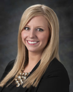 Portrait of Woman. She is looking slightly over her left shoulder and smiling. Her hair is blonde and runs down her shoulders. She is wearing a black blazer.