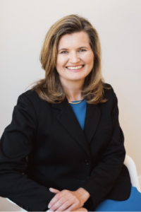 Smiling woman with shoulder length brown hair in black jacket and blue dress.