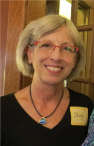 Smiling blonde woman wearing glasses and black top and a bluestone necklace.