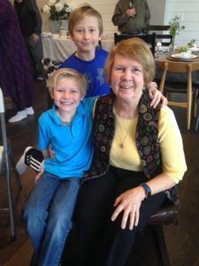 Group photo. From left to right:in front young boy seated wearing blue shirt and jeans. He is blonde haired and smiling. A sandy brown haired boy is standing behind him also smiling, also wearing blue shirt. Gail Tate is seated next to the boy to the right. She is wearing a yellow shirt, black vest, black pants. She has sandy brown hair and is smiling.